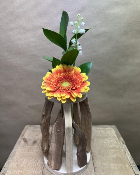 Création Originale Muguet et Bois Flotté – Fleuriste Lille Structure en bois flotté avec un gerbera orange et des brins de muguet, artisan fleuriste Openflor Lille.